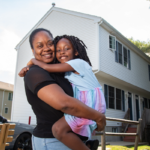 mom holding daughter as they stand outdoors in front of their home and beside their driveway