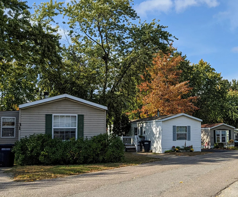 A manufactured-home community with several single-story homes lining a curved paved road and trees surrounding the neighborhood, with a few parked vehicles visible near the homes.