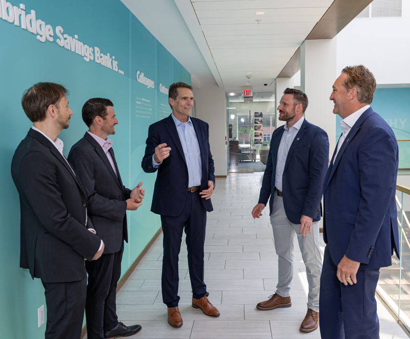 Six men wearing professional attire standing in corridor by a staircase with a glass wall. The words Cambridge Savings Bank appear on a wall behind them.