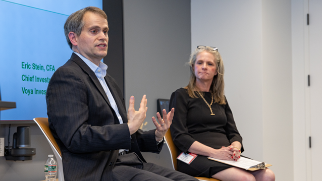 Two speakers participate in a panel discussion at the front of a meeting room while attendees listen from the audience. A presentation screen behind the speakers displays the title “Investment Manager Perspective,” and audience.