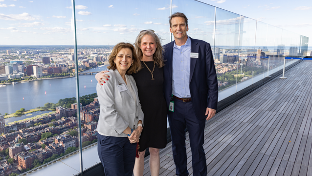 Three event attendees pose for a photo on an outdoor terrace overlooking Boston and the Charles River. A glass railing runs along the edge of the terrace, and the city skyline is visible in the background under a clear sky.