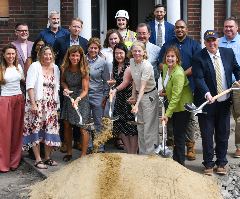 Group of men and women standing outdoors in front of a building with sand in front of them while four in the front hold shovels that they're digging into the sand.