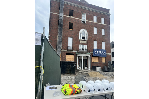 Exterior of a brick building under construction in the background with a long table consisting of construction hats and construction clothing.