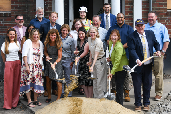 Group of men and women standing outdoors in front of a building with sand in front of them while four in the front hold shovels that they're digging into the sand.