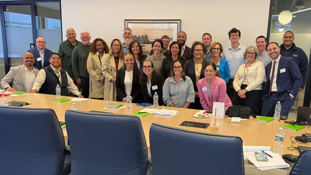 A group of approximately 25 men and women pose for a photo around a large conference room table. The room features blue conference chairs, water bottles, notebooks, and meeting materials on the table, with a framed artwork hanging on the wall in the background.
