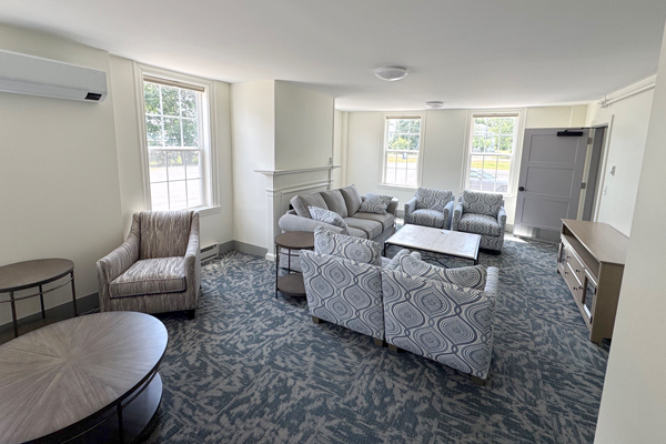 A bright light filled, carpeted common room with couches, tables, and chairs.