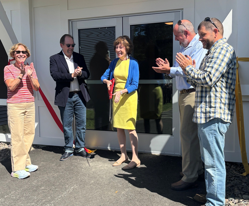 A group of two women and three men stand outside a building entrance during a ribbon-cutting ceremony as one of the women cuts a red ribbon with oversized scissors while four others stand nearby applauding.