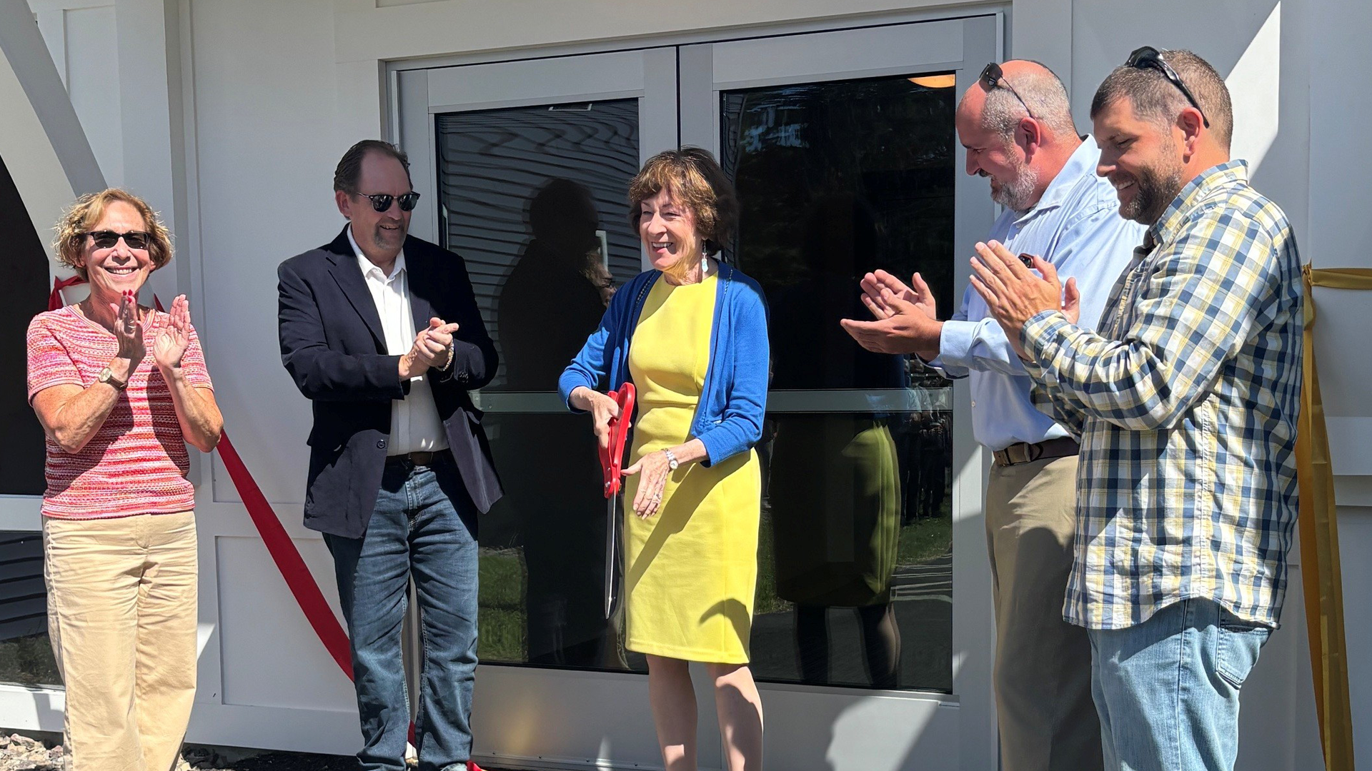 A group of two women and three men stand outside a building entrance during a ribbon-cutting ceremony as one of the women cuts a red ribbon with oversized scissors while four others stand nearby applauding.
